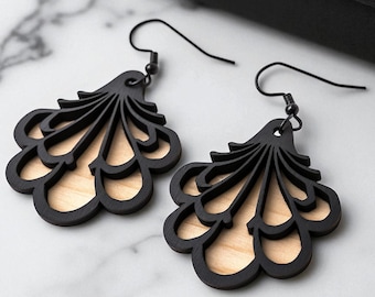 May include: A pair of black, laser-cut wooden earrings with a layered, fan-like design. The earrings have a light wood backing and black metal ear wires. The earrings are displayed on a white marble surface.