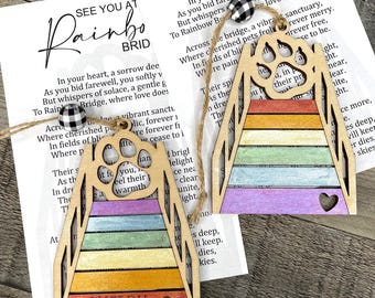 Könnte beinhalten: Zwei hölzerne Rainbow Bridge-Ornamente mit einem Pfotenabdruck und einem Herz-Ausschnitt. Jedes Ornament hat ein Regenbogenfarbenes Brückendesign und eine Juteschnur zum Aufhängen. Ein Ornament ist mit dem Namen "Shelby" personalisiert.