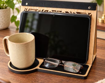 Puede incluir: Un soporte de madera para tablet con una correa superior de cuero negro y remaches plateados, un hueco circular para taza y una bandeja rectangular para gafas. Una taza de cerámica beige se asienta en el hueco y unas gafas descansan en la bandeja.