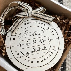 May include: A round, white wooden ornament with the text "There's no place like home 4805 for the holidays" in black. The ornament has a black and white striped ribbon and is packaged in a box with brown paper filler.