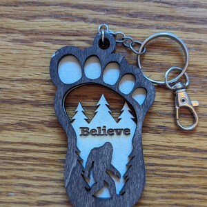 May include: A wooden keychain shaped like a Bigfoot footprint with a silhouette of Bigfoot walking through a forest inside. The keychain has the word "Believe" written on it.