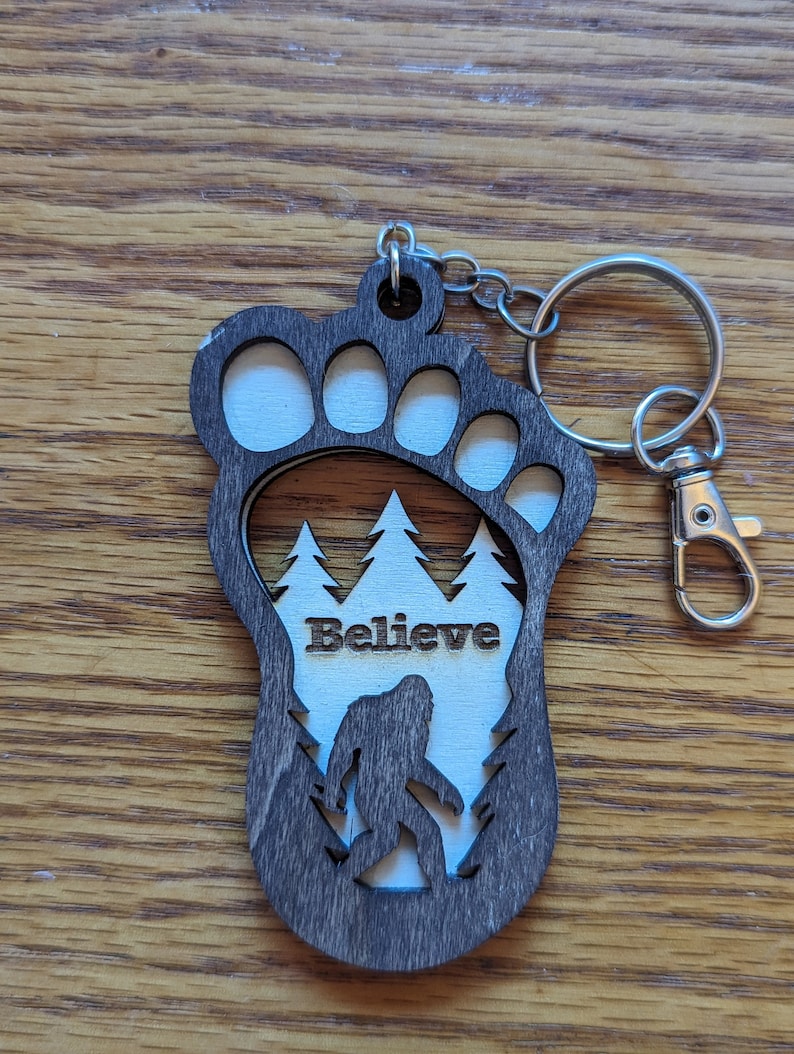 May include: A wooden keychain shaped like a Bigfoot footprint with a silhouette of Bigfoot walking through a forest inside. The keychain has the word "Believe" written on it.