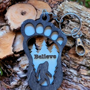 May include: A wooden keychain shaped like a Bigfoot footprint with a silhouette of Bigfoot inside. The keychain is painted black and white and has the word "Believe" written on it.