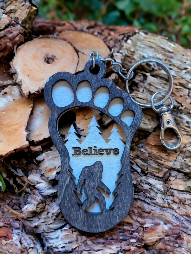 May include: A wooden keychain shaped like a Bigfoot footprint with a silhouette of Bigfoot inside. The keychain is painted black and white and has the word "Believe" written on it.