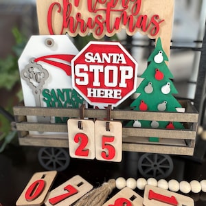 Może przedstawiać: Drewniana dekoracja w kształcie wozu z napisem Merry Christmas, znakiem Santa Stop Here, zieloną choinką z ozdobami i kluczem. Wóz zawiera drewniane płytki z liczbą 25 oraz dodatkowe płytki z cyframi 0, 7, 8 i 1 znajdujące się w pobliżu.