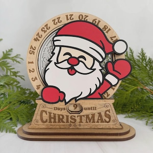 Può includere: Calendario natalizio a conto alla rovescia in legno con Babbo Natale. Il design include un quadrante circolare con numeri, una figura di Babbo Natale e la scritta "Days until Christmas". La base mostra "9" giorni rimanenti.