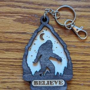 May include: A wooden keychain with a silhouette of Bigfoot walking in front of a crescent moon and stars. The keychain is dark brown with a white background and the word "BELIEVE" is engraved on the bottom.