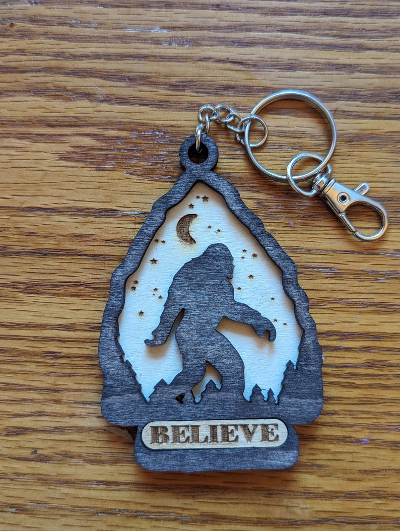 May include: A wooden keychain with a silhouette of Bigfoot walking in front of a crescent moon and stars. The keychain is dark brown with a white background and the word "BELIEVE" is engraved on the bottom.