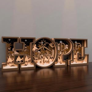 May include: Wooden "HOPE" sign with a nativity scene carved inside the "O". The letters are brown with a light wood interior, and the scene includes camels, palm trees, and a star. The sign is on a dark wooden surface.