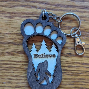 May include: A wooden keychain shaped like a Bigfoot footprint with a silhouette of Bigfoot walking through a forest inside. The keychain has the word "Believe" engraved on it.