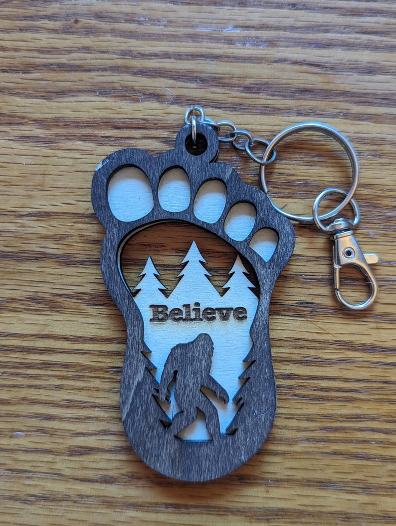 May include: A wooden keychain shaped like a Bigfoot footprint with a silhouette of Bigfoot walking through a forest inside. The keychain has the word "Believe" engraved on it.
