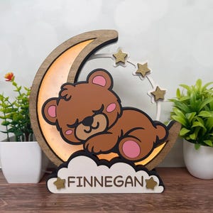 May include: A wooden night light featuring a sleeping brown bear on a crescent moon with gold stars. The name "FINNEGAN" is displayed on a cloud-shaped base. The moon has a warm, glowing light.