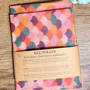 Puede incluir: Envoltorios de abeja reutilizables con un diseño colorido y estampado. Los envoltorios presentan un patrón repetido de semicírculos naranjas, rosas, morados y verdes. Una banda de papel marrón con texto envuelve los envoltorios doblados.