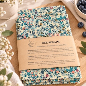 Könnte beinhalten: Ein Stapel rechteckiger Bienenwachstücher mit einem Blumenmuster in Blau, Rosa und Grün auf cremefarbenem Grund. Eine braune Papierbanderole umschließt die Mitte mit dem Text BEE-WRAPS und aufgedruckten französischen Gebrauchsanweisungen.