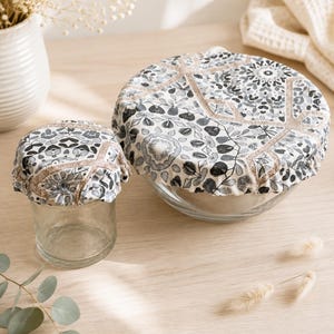 Peut inclure: Deux couvercles en tissu réutilisables avec bords élastiques, présentant un motif floral et géométrique noir, blanc et gris avec des accents beige. Une housse s'adapte à un petit bocal en verre, tandis que la plus grande couvre un bol en verre.