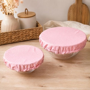 Peut inclure: Deux bols en verre ronds recouverts de housses en tissu rose à petits pois blancs. Les housses sont dotées de bords élastiqués qui s'ajustent parfaitement autour des bords des bols, assurant une fermeture sécurisée pour le stockage.