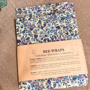 Può includere: Wrap riutilizzabili in cera d'api con un motivo floreale blu, viola e pesca su sfondo crema. Gli involucri sono piegati e hanno un'etichetta di carta marrone con testo in francese. Progettati per la conservazione degli alimenti.