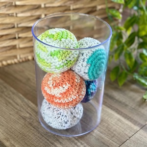 May include: A clear glass cylinder filled with six colorful, crocheted balls. The balls are various colors, including green, orange, blue, and gray. The balls are sitting on a wooden surface.