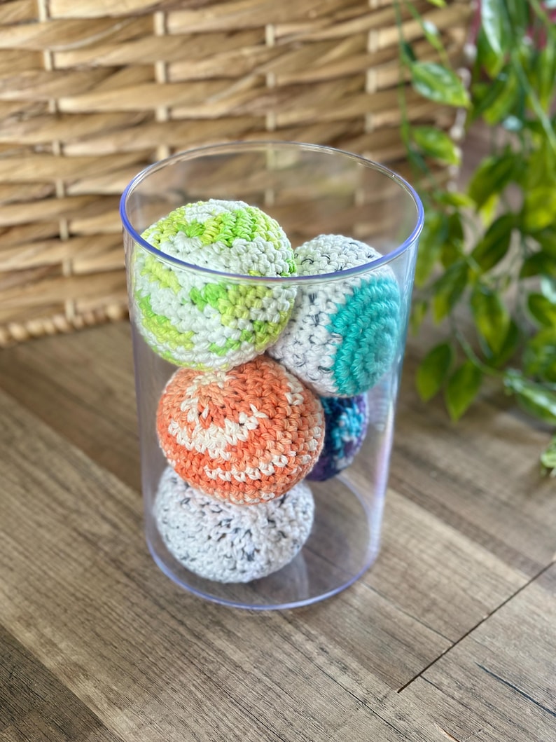 Pode incluir: Um cilindro de vidro transparente preenchido com seis bolas coloridas de croch&ecirc;. As bolas s&atilde;o de v&aacute;rias cores, incluindo verde, laranja, azul e cinza. Elas est&atilde;o em uma superf&iacute;cie de madeira.