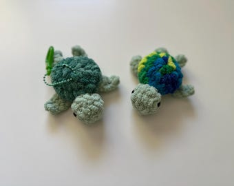 May include: Two handmade crochet turtle keychains. One is solid teal, the other is blue, green, and yellow. Both have light green heads and limbs, and black eyes. One has a green clip and chain.