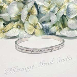 May include: Silver bangle bracelet with the word "courage" engraved around the band. The bracelet is on a white surface with white flowers in the background.