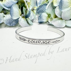 May include: A silver metal cuff bracelet with the word "COURAGE" and an arrow engraved on it. The bracelet is on a white surface with a bouquet of blue and white flowers in the background. The text "Hand Stamped by Laurie" is also visible on the surface.