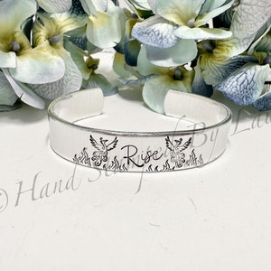 May include: A silver metal cuff bracelet with the word "Rise" engraved on it. The bracelet is decorated with two phoenixes rising from flames.