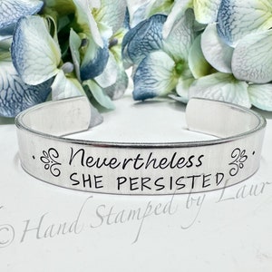 May include: Silver bracelet with the inscription "Nevertheless, She Persisted" and a small floral design on either side. The bracelet is hand-stamped by Laurie.