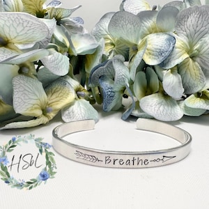 May include: Silver bracelet with an arrow and the word "Breathe" engraved on it. The bracelet is on a white surface with blue and white flowers.