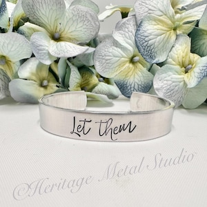 May include: Silver metal cuff bracelet with the words "Let them" engraved in black lettering.