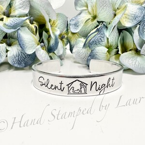 May include: A silver metal cuff bracelet with the words "Silent Night" and a depiction of the nativity scene. The bracelet is hand-stamped with the text "Hand Stamped by Laurie".