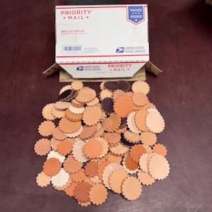 以下が含まれることがあります： さまざまな茶色とタンの色合いの、小さなスカラップエッジの革製円の山。円は暗い表面に散らばっており、背景には部分的に見えるPriority Mailの箱があります。