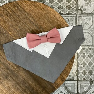 May include: A gray and white dog bandana with a pink bow tie. The bandana has a white collar and three buttons.