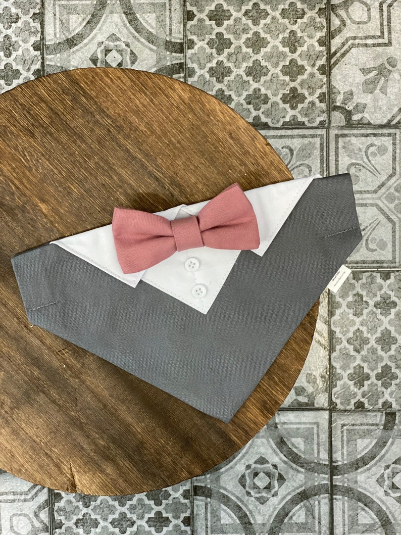 May include: A gray and white dog bandana with a pink bow tie. The bandana has a white collar and three buttons.