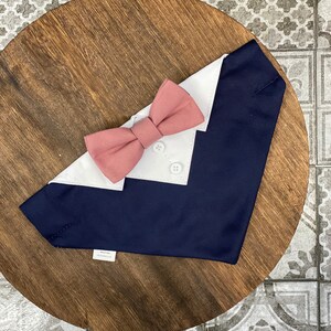 Puede incluir: Un bandana para perro azul marino y blanco con una pajarita rosa. El bandana tiene un cuello blanco y dos botones blancos.