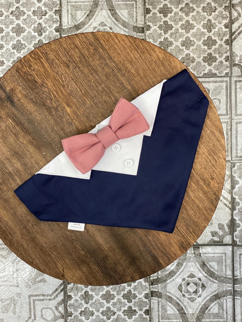 Puede incluir: Un bandana para perro azul marino y blanco con una pajarita rosa. El bandana tiene un cuello blanco y dos botones blancos.