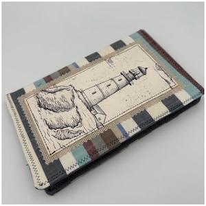 May include: A fabric wallet with a lighthouse illustration. The wallet features a rectangular design with a central image of a lighthouse on a rocky cliff. The background has a striped pattern in shades of gray, blue, and brown.