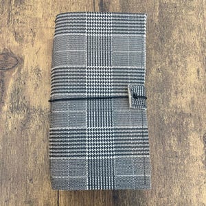 May include: A gray and white plaid journal with a black elastic closure and a silver-tone metal clasp. The journal is rectangular and features a classic houndstooth pattern. The cover is made of a textured fabric.
