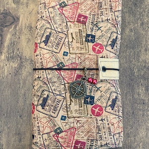 May include: A travel journal with a beige cover featuring a collage of travel stamps in red, blue, and black. The journal is secured with a black elastic band and a decorative charm. The stamps include city names and dates.