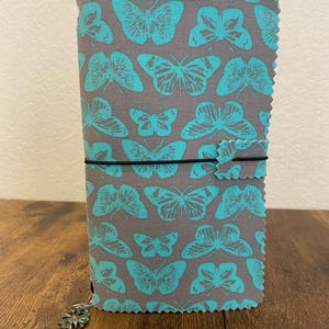 May include: A gray journal cover with a teal butterfly pattern. The journal is secured with a black elastic band and features a decorative charm. The edges of the cover have a decorative, scalloped edge.