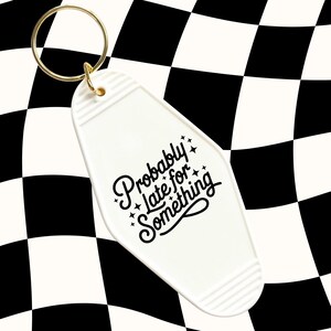 May include: A white motel-style keychain with a gold ring. The keychain has the phrase "Probably late for Something" in black script. The background is a black and white checkered pattern.