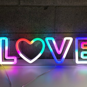 May include: A neon sign displaying the word "LOVE" in vibrant, multi-coloured lights. The letters are outlined in white and illuminated with a spectrum of colours, including pink, green, blue, and yellow. The heart-shaped "O" adds a romantic touch.
