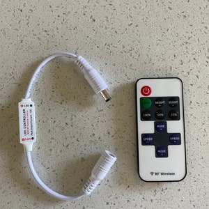 May include: A white LED controller with a white wire and a white remote control. The controller has a red LED light and the text "LED CONTROLLER" and "With RF Remote Control". The remote control has buttons for "LIGHT", "BRIGHT", "MODE", and "SPEED".