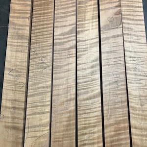 Könnte beinhalten: Sechs Holzbretter in hellem Braun mit einem wellenförmigen Maserungsmuster. Die Bretter weisen handschriftliche Markierungen auf, darunter "215 Test". Das Holz ist unbehandelt und für Holzbearbeitungsprojekte geeignet. Ideal für Möbel oder andere Holzartikel.