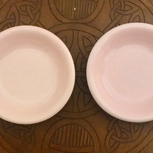 Puede incluir: Dos cuencos pequeños y redondos de color rosa claro. Los cuencos tienen un diseño simple y poco profundo y están colocados sobre un fondo marrón con dibujos. Los cuencos probablemente son para servir pequeñas porciones de comida o para fines decorativos.