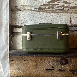 Può includere: Valigia vintage verde oliva con chiusure e manico in metallo argentato. La piccola valigia da viaggio è strutturata e poggia su una superficie di legno. La valigia misura circa 30 cm di lunghezza.
