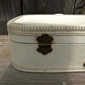 May include: An off-white vintage suitcase with a textured exterior and a decorative woven trim. The suitcase features a tarnished, ornate metal clasp and hinges. The rounded edges and overall design suggest a classic travel accessory.