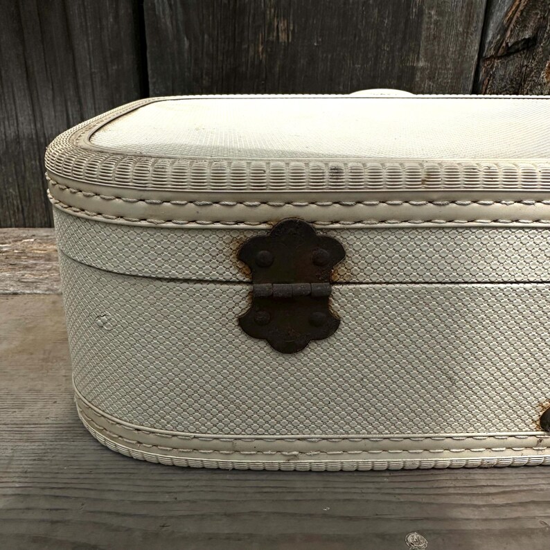 May include: An off-white vintage suitcase with a textured exterior and a decorative woven trim. The suitcase features a tarnished, ornate metal clasp and hinges. The rounded edges and overall design suggest a classic travel accessory.