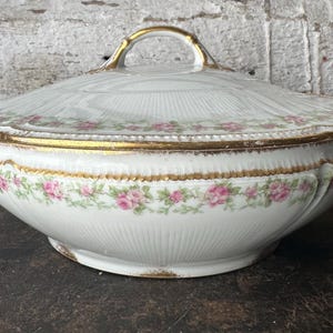 May include: White porcelain serving dish with lid, featuring a gold trim and handle. The dish is decorated with a floral pattern of pink roses and green leaves. The dish has a ribbed texture and is likely vintage.