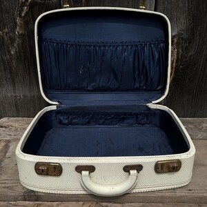 May include: Open vintage cream-colored suitcase with a navy blue interior. The suitcase has a white handle, brass-colored latches, and a textured exterior. The interior features pockets and a lined base, suggesting it's designed for travel or storage.